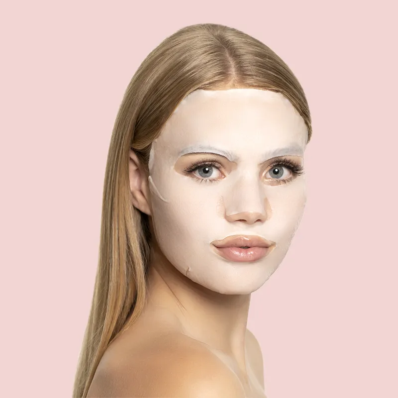 Frau mit blonder, glatter Frisur trägt eine weiße Gesichtsmaske vor einem rosa Hintergrund.