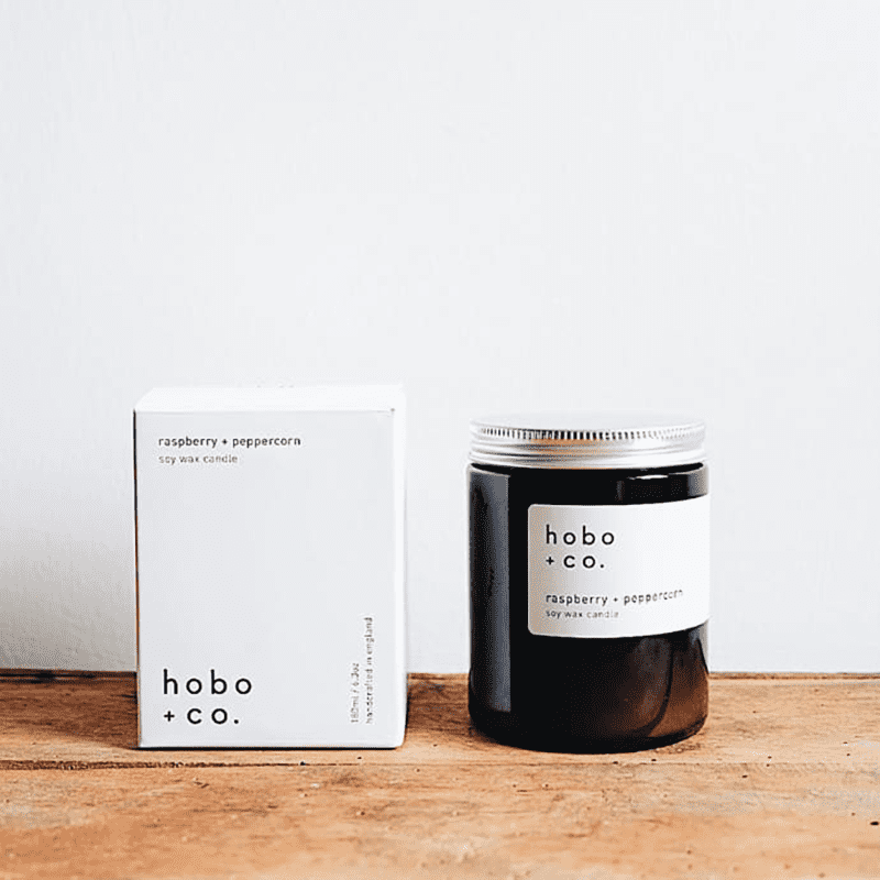 hobo-co-raspberry-peppercorn-essential-oil-candle-180ml_optimized Hobo + Co. Sojawachskerze mit Himbeer-Pfefferkorn-Duft, 180ml, neben der Verpackung auf einem Holztisch.