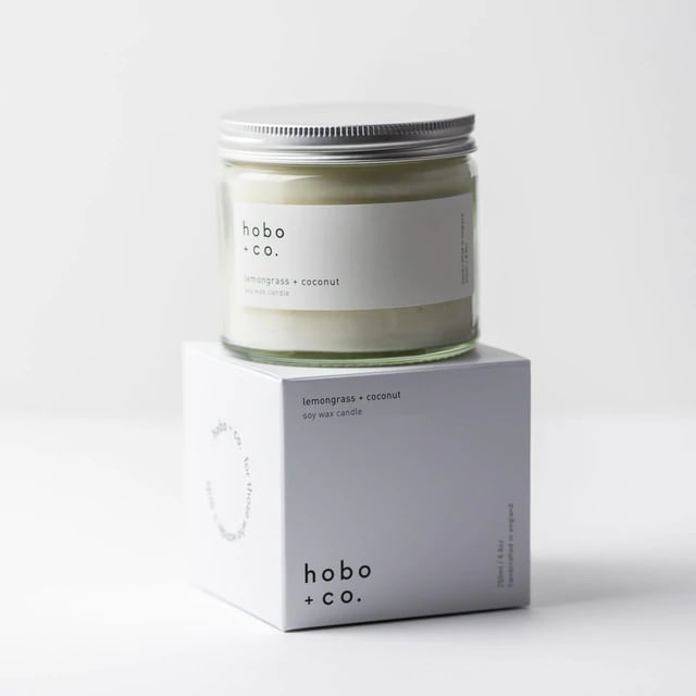 Kerze im Glas mit Zitronengras-Kokosduft von hobo + co. auf einer weißen Verpackung.