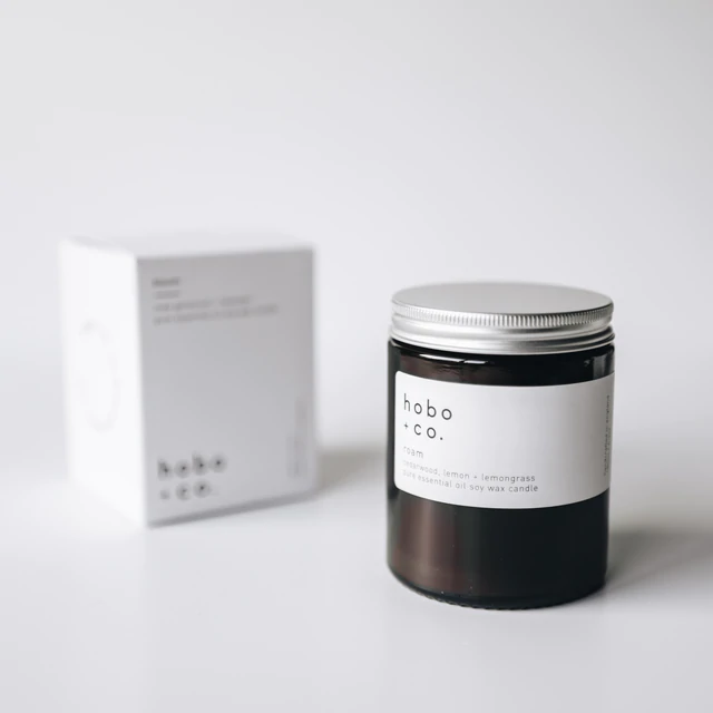 Eine schwarze Duftkerze von hobo + co. mit der Aufschrift 'Cedarwood, Lemon + Lemongrass' steht vor einer weißen Verpackung.