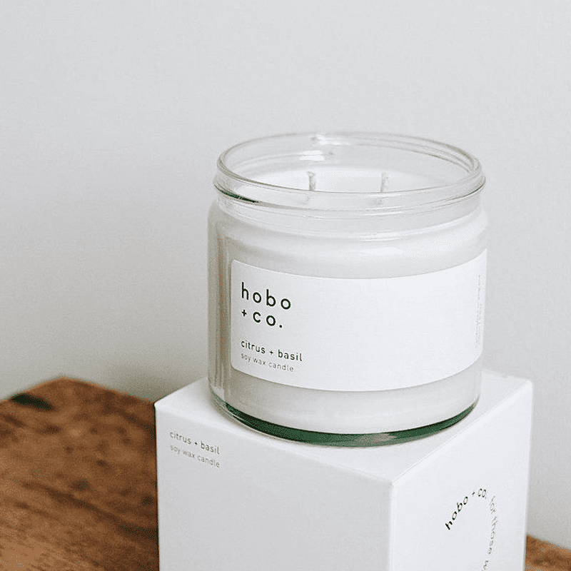 hobo-co-citrus-basil-essential-oil-candle-250ml Hobo + Co. Zitrus-Basilikum Sojawachskerze in einem Glasbehälter auf einer weißen Box.