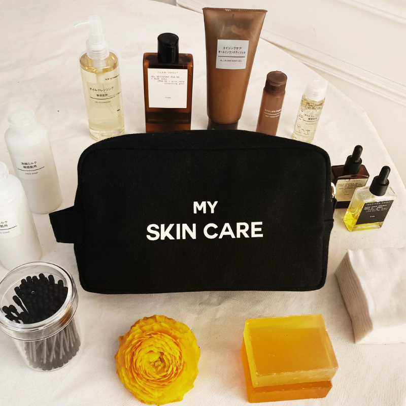 Schwarze Kosmetiktasche mit der Aufschrift 'MY SKIN CARE' umgeben von Hautpflegeprodukten auf einem weißen Hintergrund.