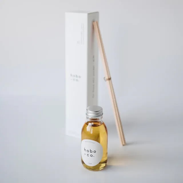 Kleine Flasche mit Duftöl von hobo + co. und Holzstäbchen vor einer weißen Verpackung auf einem weißen Hintergrund.